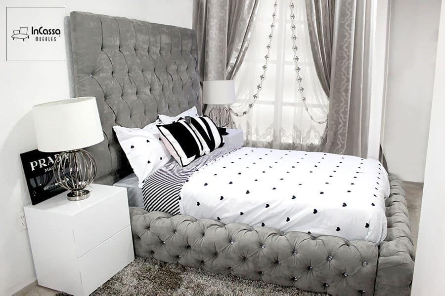 Esta imagen muestra una cama con un diseño elegante y moderno, destacando su cabecera y base tapizadas con capitoné en tela de suede gris perla, acompañadas por dos buroes blancos. La estructura es robusta, proporcionando un aspecto lujoso y acogedor a la habitación.