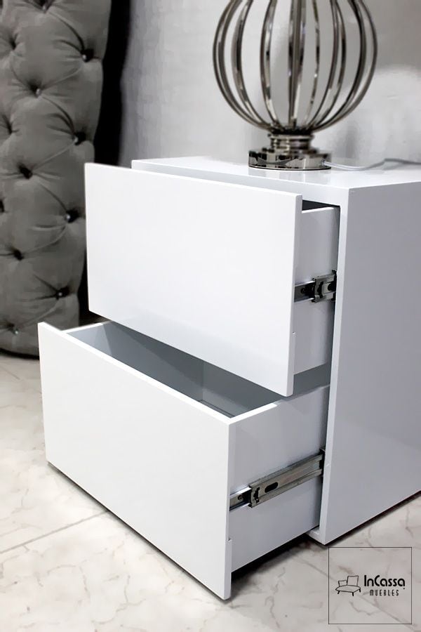 La foto muestra un buroe blanco con un diseño moderno y minimalista. Cuenta con dos cajones con rieles metálicos para apertura suave, ideales para almacenamiento adicional en el dormitorio. El acabado brillante le da un toque elegante y contemporáneo.