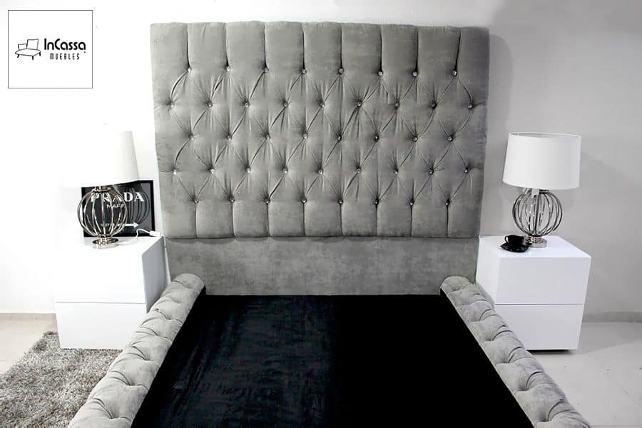 La imagen muestra una cama con una cabecera alta y tapizada en suede gris perla, con un acabado de capitoné que incluye botones decorativos. La base de la cama también presenta un diseño capitoné, añadiendo un toque de lujo y sofisticación. El conjunto está acompañado por dos buroes blancos, creando un contraste moderno y elegante.