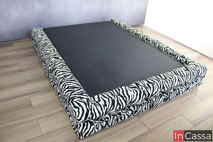 Vemos la base desde una toma lateral que nos da una vista más completa del mueble, en está foto la base se presenta con un tapizado en lino estilo cebra con acabado capitoneado, y se encuentra instalada frente a una pared gris y sobre un piso de madera.