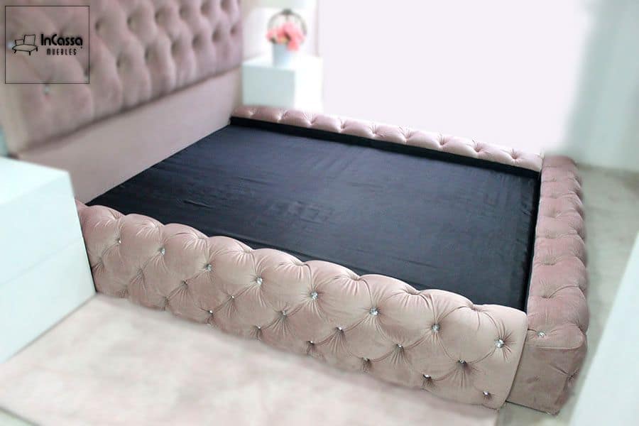 La base es presentada junto a una cabecera tapizada en suede rosa palo con acabado capitoneado al igual que la base, Además pueden notarse dos buroes blancos acompañando ambos muebles.