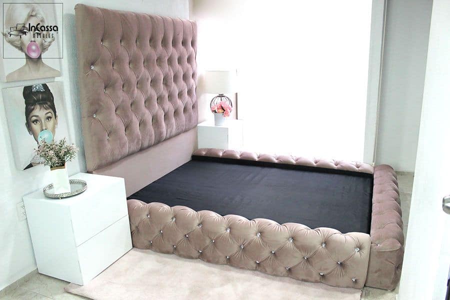La imagen presenta una elegante cama con una cabecera alta y tapizada en suede rosa palo con un acabado capitoneado, adornado con botones decorativos. La base de la cama también sigue el mismo diseño capitoné, creando una apariencia cohesiva y lujosa. El ambiente está complementado con buroes blancos y una decoración de arte pop en la pared, que añade un toque moderno y artístico al espacio.