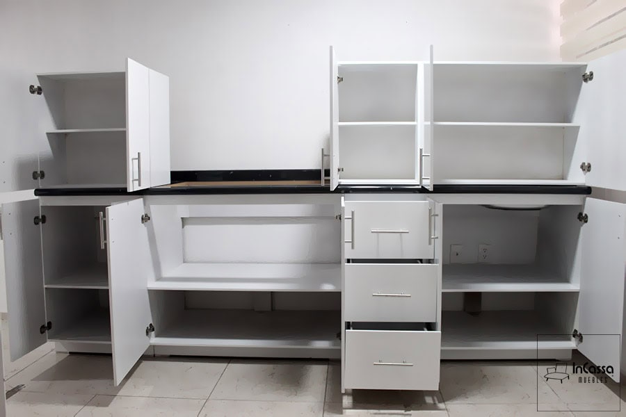 Cocina modular abierta con gabinetes blancos y cubierta de formaica negra, mostrando amplio espacio de almacenamiento. Diseño funcional con estantes ajustables y jaladeras de barra metálicas, ideal para optimizar el orden en el hogar.