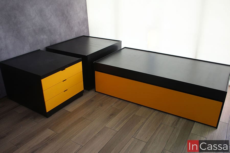 Foto de la trilitera de acabado chocolate mate, en la que se muestra con sus módulos levemente separados, siendo un total de tres entre los cuales está una base doble individual, un módulo central y un módulo con cajones amarillos, sirviendo estos dos últimos para formar la base superior.