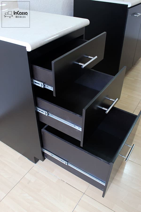 La imagen muestra una toma lateral que destaca tres cajones abiertos del módulo inferior de cocina. Cada uno cuenta con frentes en color chocolate mate, correderas metálicas de extensión y jaladeras rectas de acero inoxidable. La cubierta de formaica superior en tono granito de arena con acabado brillante aporta contraste al diseño. Esta estructura es ideal para maximizar el almacenamiento con estilo moderno y funcional.