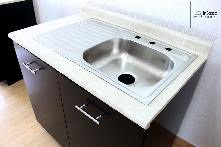 Esta foto presenta un enfoque en ángulo que resalta el módulo inferior con puertas en color chocolate mate y jaladeras metálicas. La cubierta de formaica en tono granito de arena incorpora una tarja sencilla de acero inoxidable con escurridor lateral y perforaciones para instalación de mezcladora. Un diseño práctico y moderno, ideal para cocinas funcionales con estilo contemporáneo.