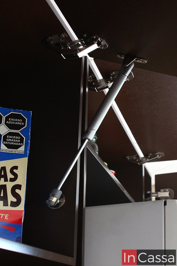 Este detalle muestra el pistón neumático que permite que las puertas de los gabinetes superiores de color tabaco se mantengan abiertas de forma suave y segura, evitando golpes o cierres abruptos.