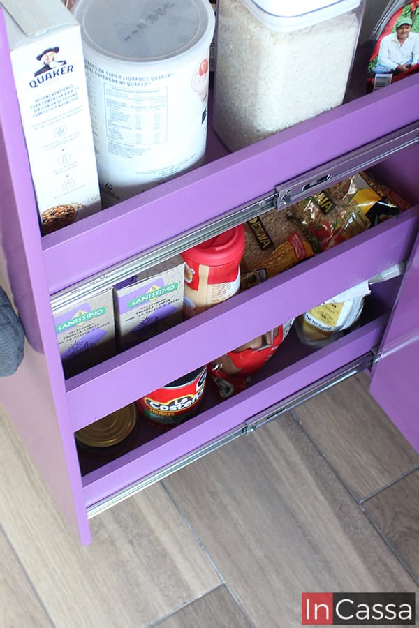 La foto presenta el especiero extraíble del mueble carnicero en color uva completo, lleno de diversos productos básicos de cocina como avena, arroz y conservas. Este diseño práctico facilita el acceso y la organización de los ingredientes diarios en un espacio compacto y funcional.