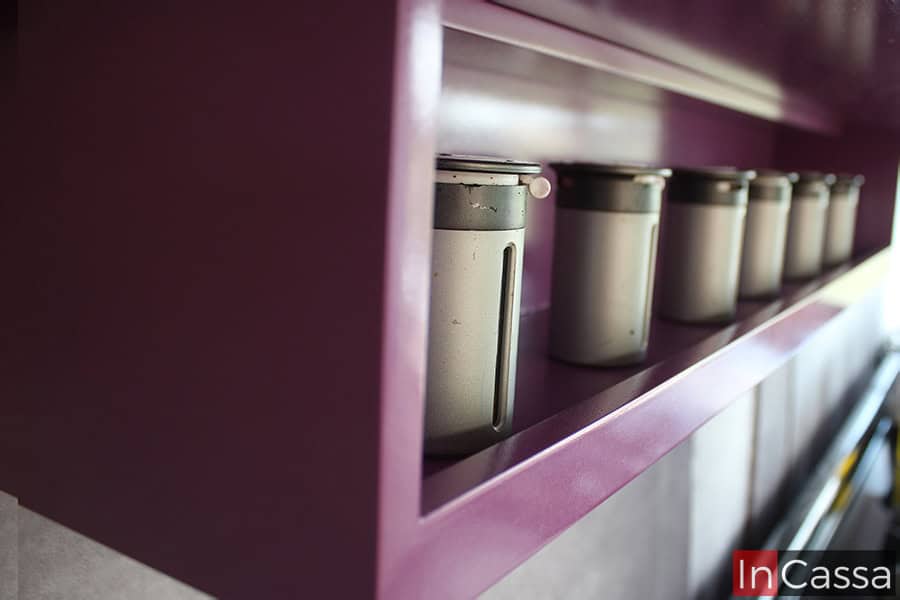 La foto presenta un acercamiento a un especiero integrado en la alacena superior de color uva completo, donde se alinean varios contenedores metálicos. Este diseño práctico y elegante permite tener los condimentos al alcance de la mano, optimizando el espacio en la cocina.