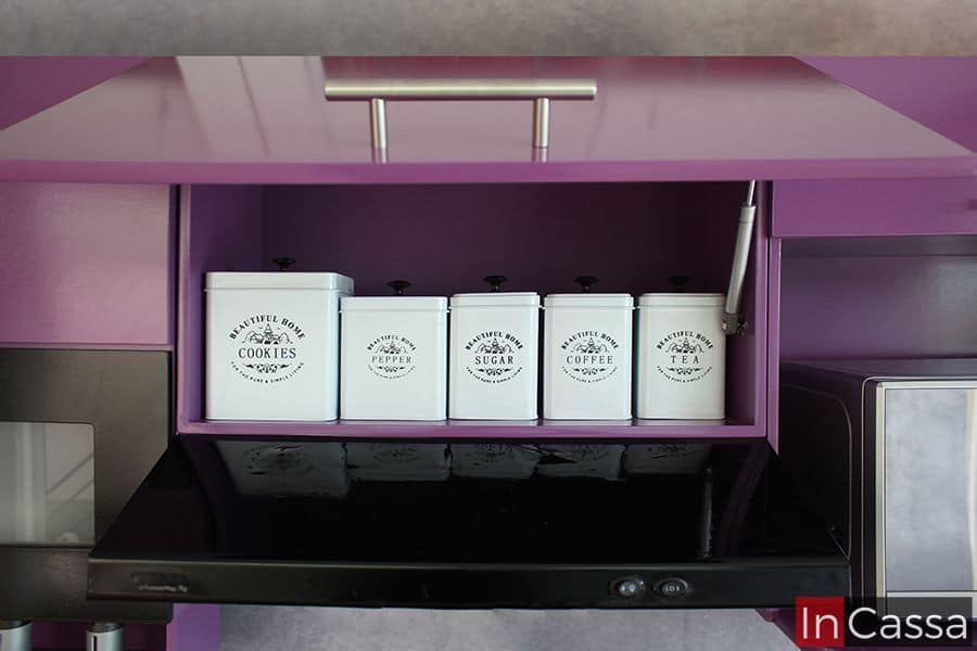 Una fotografía del módulo superior sobre campana en color uva, con pistones neumáticos para la apretura y cierra de su puerta. Se presenta con botes metálicos etiquetados para galletas, pimienta, azúcar, café y té. Este espacio es perfecto para mantener los ingredientes esenciales siempre a mano.