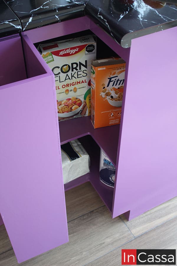 La toma se enfoca en el mueble esquinero inferior, en el que se puede encontrar dos compartimentos adicionales ocultos por una puerta en forma de L. Estos espacios internos conservan el mismo color uva que puede verse en toda la cocina.