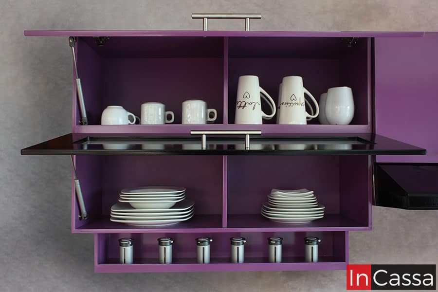 Foto de la alacena superior de la cocina, presentada con sus puertas abiertas revelando 4 compartimentos en su interior los cuales mantienen el mismo color uva que puede verse en toda la cocina. Las dos puertas del módulo cuentan con un sistema de apertura y cierre vertical gracias a pistones neumáticos, además la alacena cuenta con un pequeño espacio en su parte inferior para colocar especias.