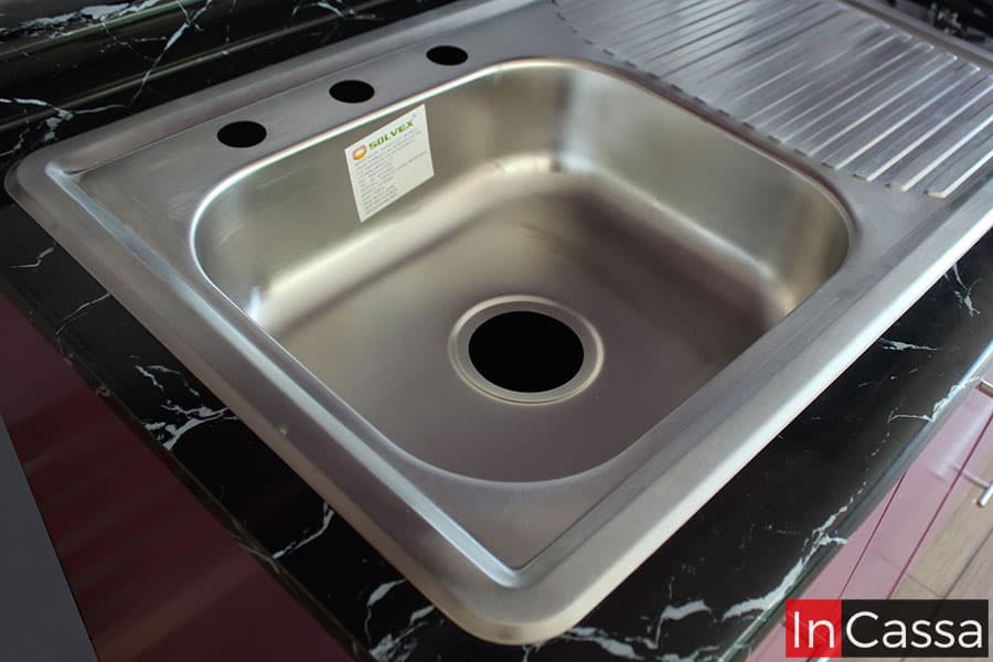 La foto se enfoca en la tarja de acero inoxidable incluida en la cocina, en la que se puede encontrar una tina sencilla en su lado izquierdo y un escurridor en el derecho. La foto además también permite apreciar parte de la cubierta de formaica de estilo mármol negro incluida en el módulo.