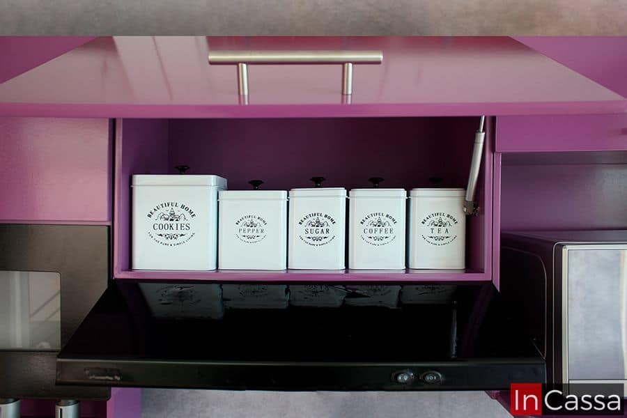 Una toma frontal de los módulos superiores de la cocina, en la que se resalta el mueble sobre campana, el cuál se muestra con su puerta abierta revelando un amplio compartimento y dejando a la vista sus pistones neumáticos integrados para la apertura y cierre vertical de la puerta. El módulo es de color uva en su totalidad y su puerta además cuenta con una jaladera metálica de barra T.