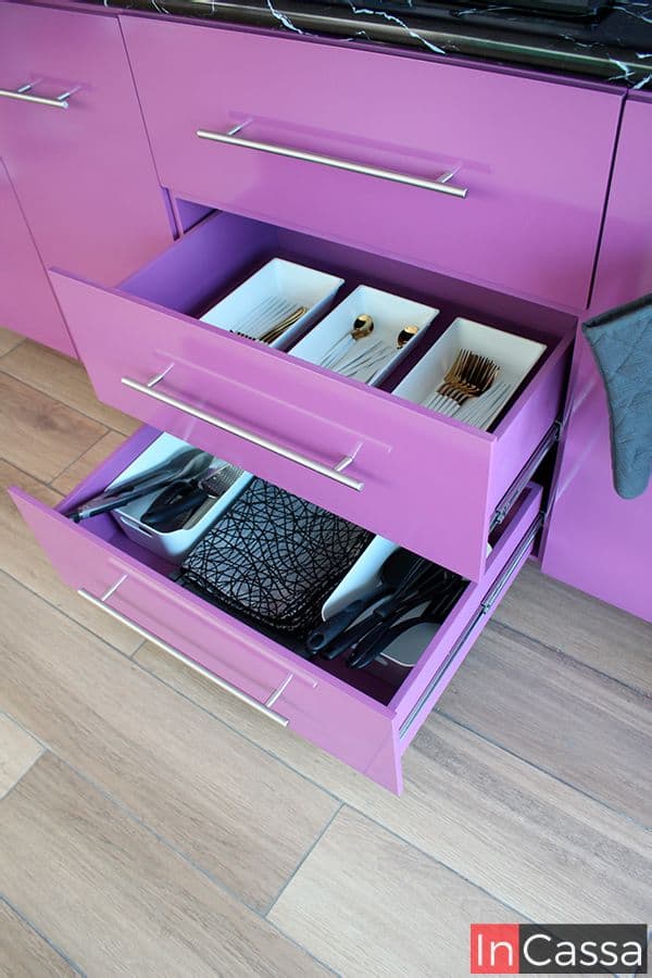 La imagen muestra los cajones integrados al módulo inferior de la cocina, estos son presentados abiertos dejando a la vista su capacidad interna. Se puede apreciar que la totalidad de la estructura de cada cajón conserva el mismo color uva presente en toda la cocina; además también es posible ver el sistema de rieles que permite la apertura y cierre de cada compartimento.