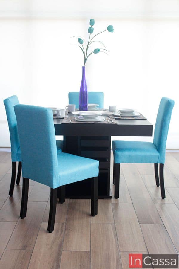 Una foto frontal del comedor presentado junto a una venta completa y un piso de madera, la iluminación que proporciona la ventana resalta la pintura chocolate mate de la mesa al igual que el tapizado en pliana aqua de las cuatro sillas incluidas en el comedor.