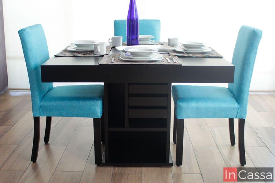 Podemos observar la estructura completa de la mesa del comedor pintada en su totalidad en color chocolate, el pedestal queda a la vista por lo que es posible ver los compartimentos con los que cuenta. Además también tenemos una vista lateral completa de dos sillas que acompañan al comedor, así como el respaldo de una tercera, todas tapizadas en pliana color aqua. El mueble está presentado junto a una ventana completa que sirve como un estupendo fondo.