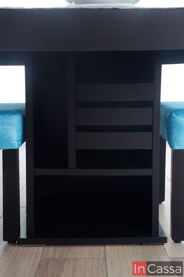 Vemos una toma completa del pedestal de la mesa, el cuál tiene un diseño minimalista con 3 compartimentos de diferentes tamaños distribuidos en la estructura, además de 3 cajones distribuidos con un poco de espacio entre ellos haciendo posible la apertura y cierre sin necesidad de jaladeras. El pedestal tiene un acabado chocolate mate al igual que el resto del comedor.