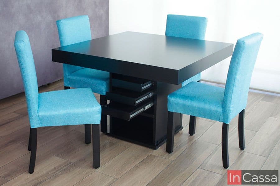 En esta fotografía se presenta una toma completa del comedor color chocolate con 4 sillas tapizadas en pliana aqua, donde es posible observar los 3 cajones incorporados en el pedestal junto con parte de su mecanismo de apertura y cierre. El conjunto es presentado junto a una venta en pantalla completa que, junto con un piso de madera, proporcionan un fondo que resalta los acabados del comedor.