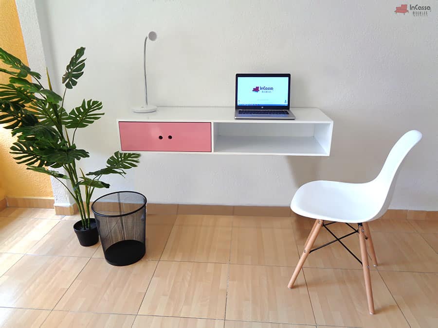 La imagen muestra un escritorio flotante en blanco con cajón rosa y compartimento abierto. Su diseño moderno y compacto se complementa con una silla blanca, lámpara y laptop, creando un ambiente funcional y elegante, ideal para oficinas o estudios en el hogar.