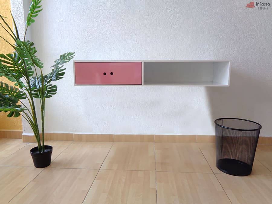 La foto muestra un escritorio flotante en acabado blanco con un cajón rosa de tiradores perforados y un compartimento abierto. Su estructura minimalista y funcional es ideal para mantener el espacio organizado con un estilo elegante y versátil.