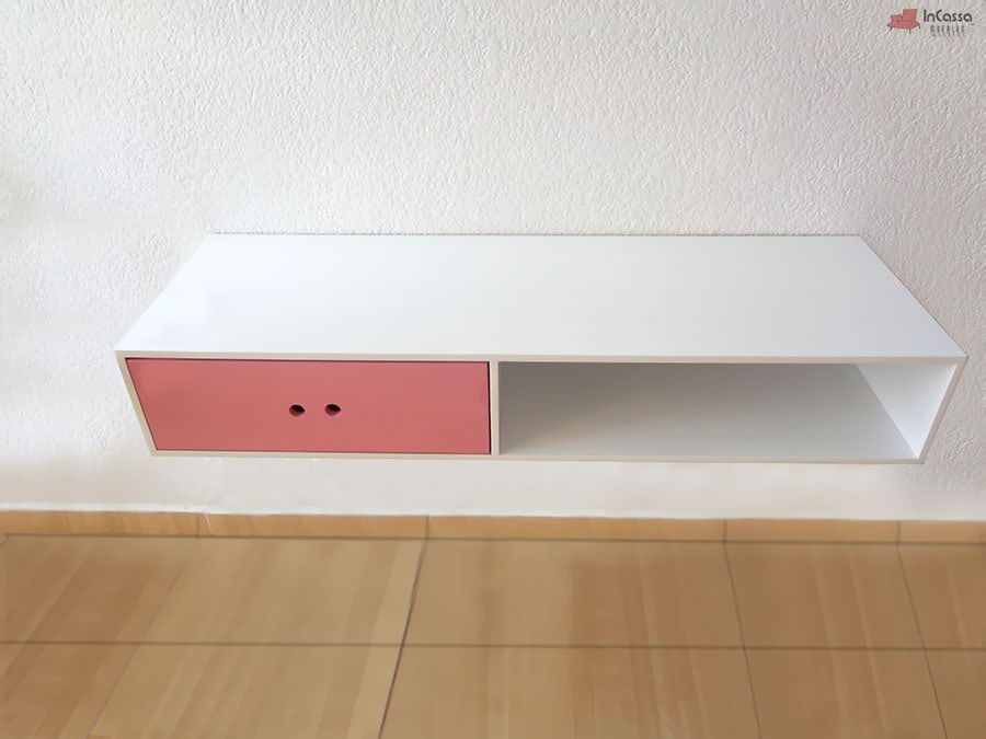 La foto muestra un mueble flotante moderno con estructura en color blanco y un cajón rosa con perforaciones como tiradores. Su diseño minimalista incluye un compartimento abierto, ideal para almacenamiento práctico en cualquier espacio.