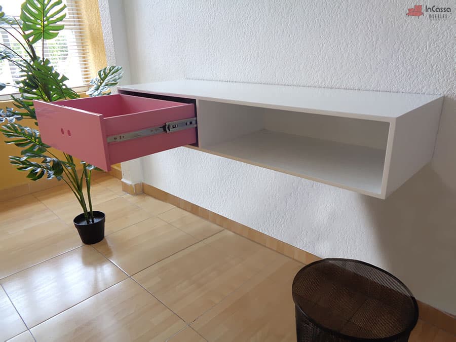Se presenta un escritorio flotante en blanco con un cajón rosa extendido, revelando sus correderas metálicas de extensión. Su diseño moderno y funcional incluye un compartimento abierto, ideal para organizar documentos o accesorios, aportando estilo y practicidad a cualquier espacio.