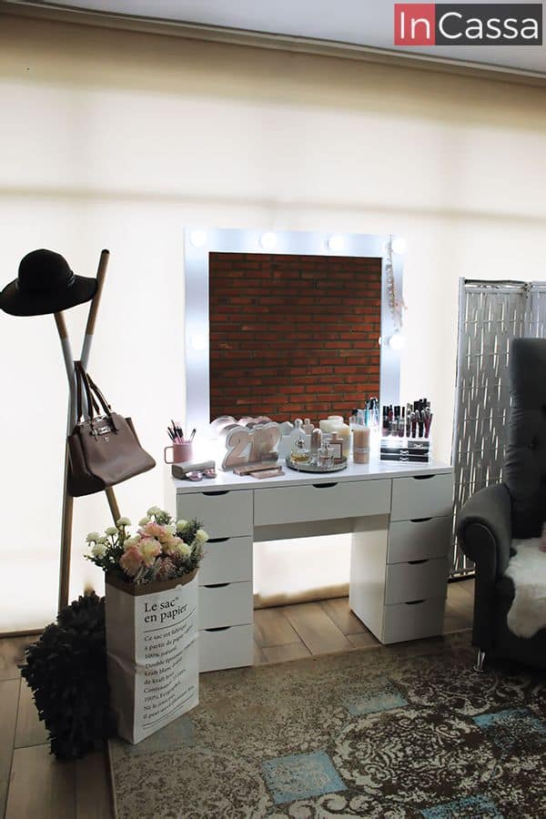 Se nos presenta el tocador blanco tipo vanity en una extravagante habitación con ventana completa y piso de madera. Gracias a las luces proporcionadas por los múltiples focos incorporados en el espejo, podemos notar que esta versión cuenta con una gruesa cubierta de MDF chileno.