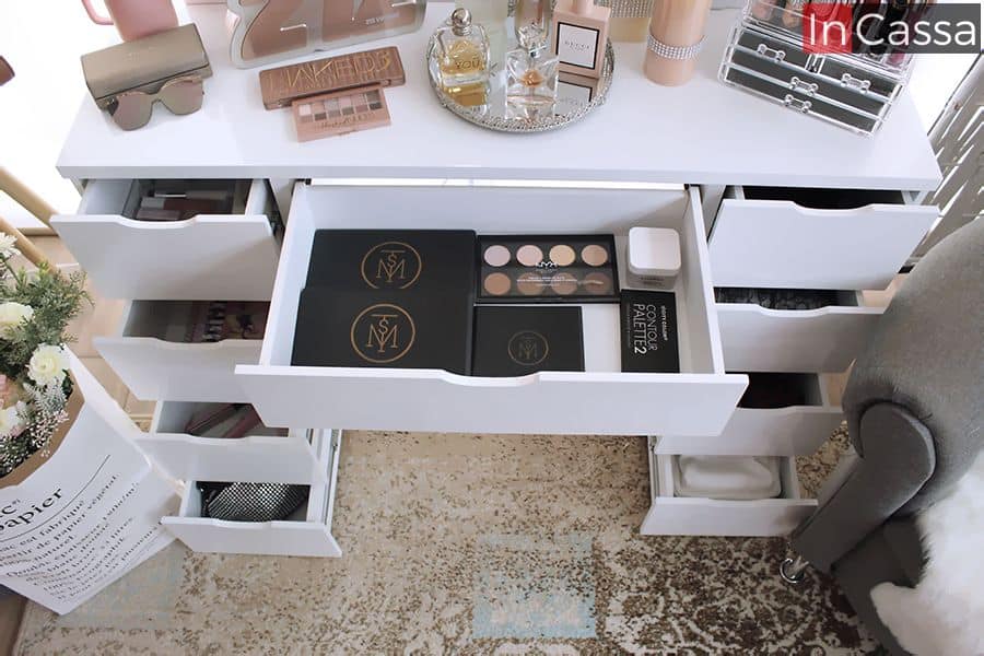 La foto nos proporciona una mejor vista de la cubierta de MDF presente en está versión del tocador así como de la estructura del cajón central y una vista parcial de la de los cajones laterales; todos pintados en un brillante color blanco al igual que todo el mueble. Se hace uso de varios utensilios y maquillajes para decorar y ejemplificar la longitud de la cubierta y la profundidad de los cajones.
