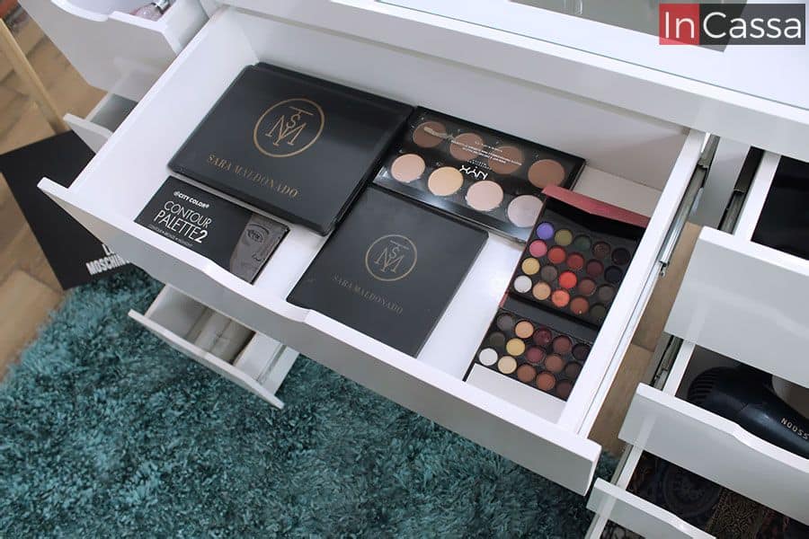 Podemos apreciar el interior del cajón central incluido en el tocador, el cual es visiblemente más ancho y espacioso que los cajones laterales, en este se almacenan varios juegos de maquillaje con los cuales podemos determinar su profundidad y extensión. Al igual que en el resto de los cajones y en la propia estructura del tocador, el cajón central presenta un tono blanco completo.