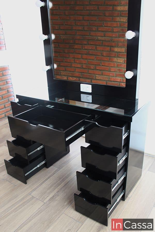 Vemos el tocador vanity en color negro junto a una ventana de pantalla completa, se nos presentan abiertos todos los compartimentos incorporados en el tocador, por lo que es posible ver que su estructura entera conserva el mismo tono negro que el tocador, además de que es posible notar parte de su sistema de apertura y cierre guiado por rieles.