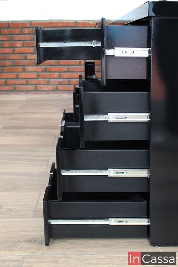 Una vista lateral del tocador en color negro, en la cual resalta sobre todo la estructura de los cajones así como su sistema de rieles metálicos incorporado. La construcción de cada cajón tiene el mismo color negro presente en toda la estructura del tocador.