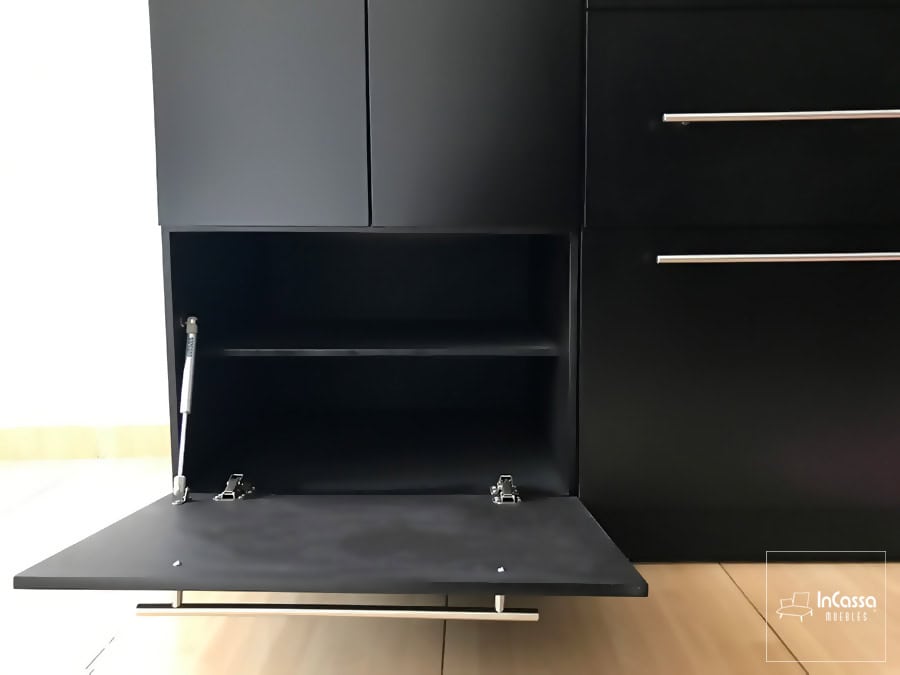 Está imagen muestra uno de los compartimentos inferiores del clóset, el cuál cuenta con un pistón neumático para la apertura y cierre de su puerta de manera vertical. El espacio es dividido en dos compartimentos gracias a un entrepaño, toda esta parte interior mantiene el mismo tono chocolate que puede apreciarse en el resto del clóset.