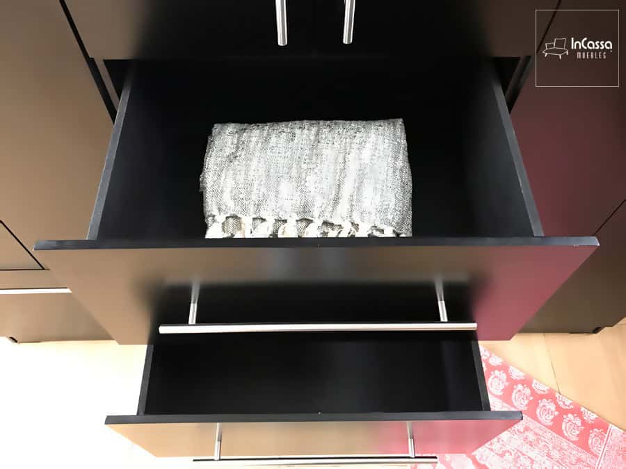 Se nos muestra una toma superior de los cajones del clóset, los cuales se encuentran abiertos dejándonos ver su interior, siendo este bastante profundo y que además mantiene el mismo tono chocolate presente en la totalidad del mueble. Igualmente se tiene una vista cercana de las jaladeras de barra T integrada a cada cajón.