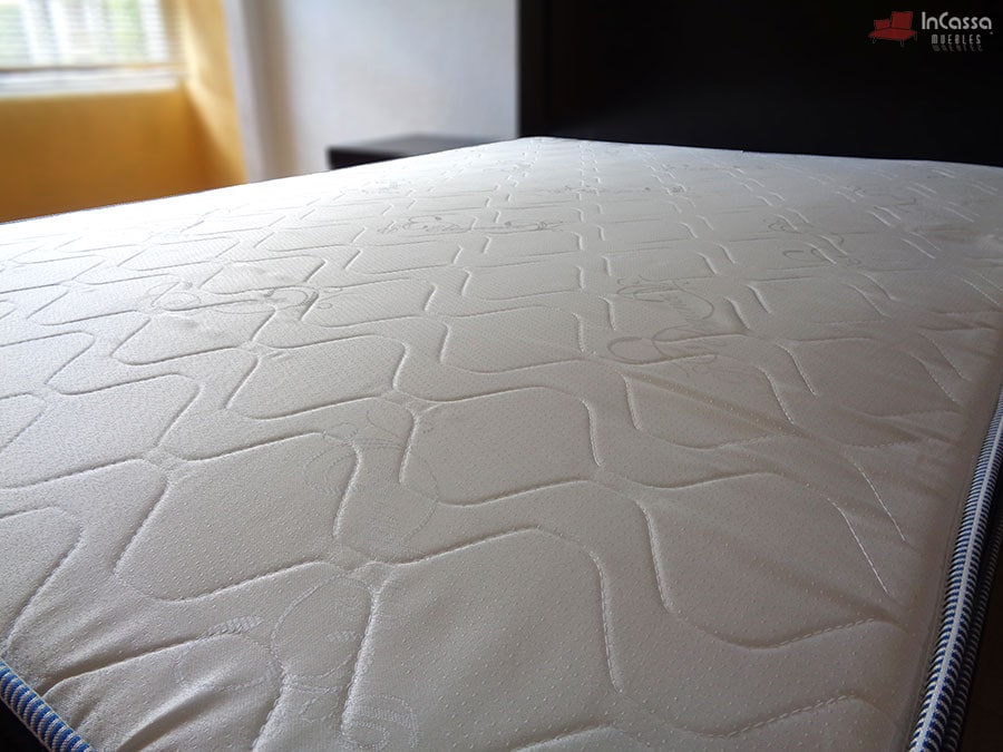 La foto enfoca el acolchonado del colchón, el cuál presenta un bordado con patrón de lineas curvas y un tono hueso, además es posible apreciar la base y buroe de color chocolate que acompañan la presentación del colchón.
