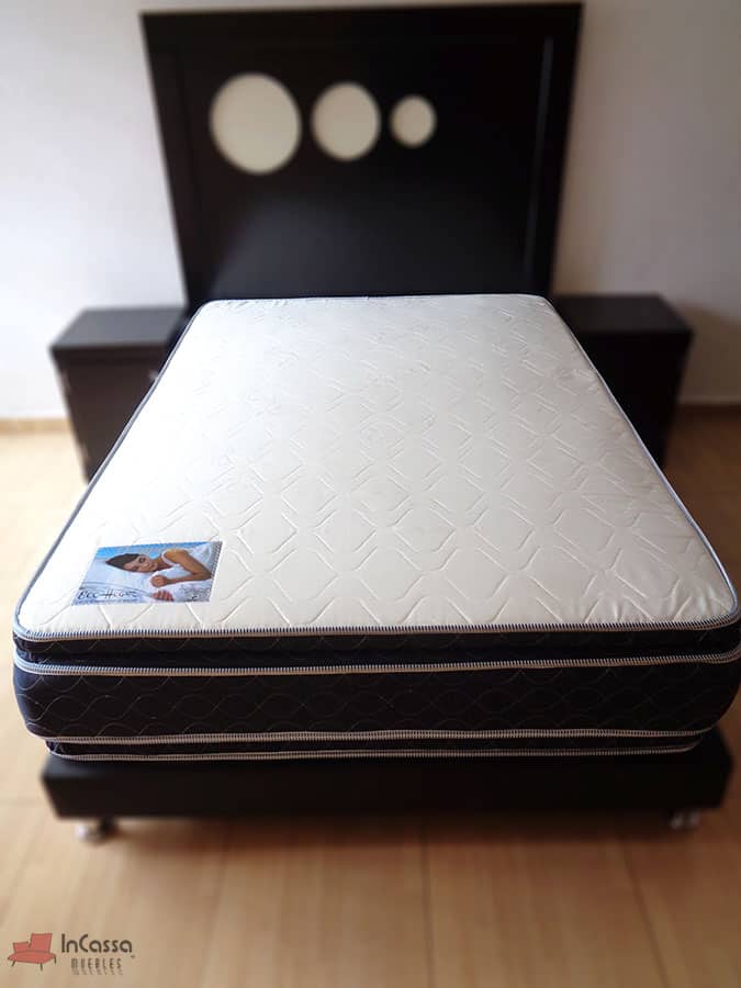 La imagen muestra una toma completa del acolchonado hueso del colchón, en el cual es más notable su bordado con patrón de lineas curvas y su etiqueta de presentación. Además esta vez podemos observar más a detalle la cabecera y buroes que conforman la recámara chocolate en la que se presenta el colchón.