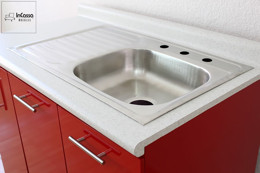 La foto muestra una toma en ángulo superior de la tarja con escurridor de acero inoxidable, perfectamente integrada en una cubierta de formaica estilo sal y pimienta. Los módulos inferiores lucen un acabado en rojo vivo brillante y jaladeras metálicas cilíndricas, aportando un contraste moderno y elegante.