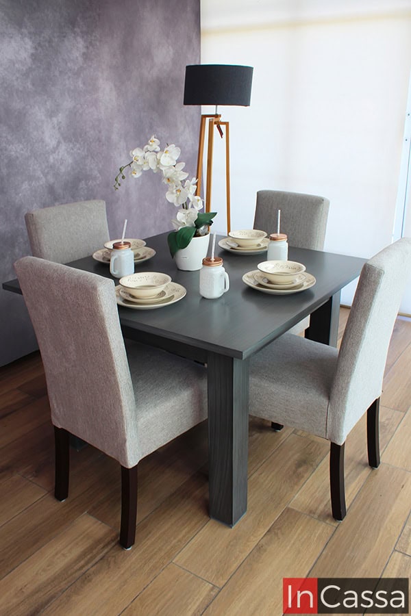 La imagen muestra un comedor de cuatro sillas tapizadas en pliana gris perla alrededor de una mesa cuadrada en acabado gris cenizo oscuro. La lámpara de pie y los accesorios decorativos completan un espacio moderno y acogedor.