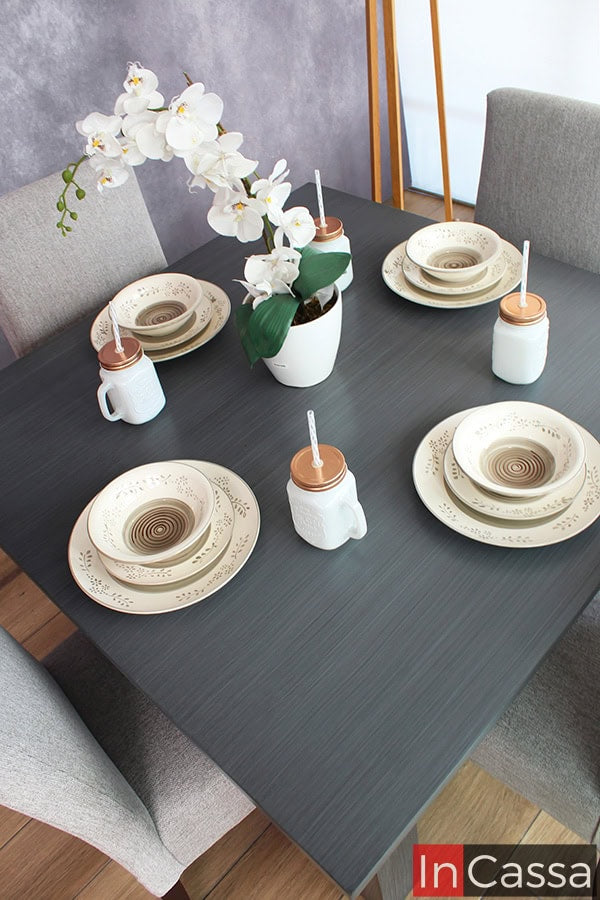 Se presenta la mesa en acabado gris cenizo oscuro de comedor decorada con vajilla y un centro de mesa con orquídeas blancas, creando un ambiente elegante y acogedor, ideal para cenas formales y casuales.