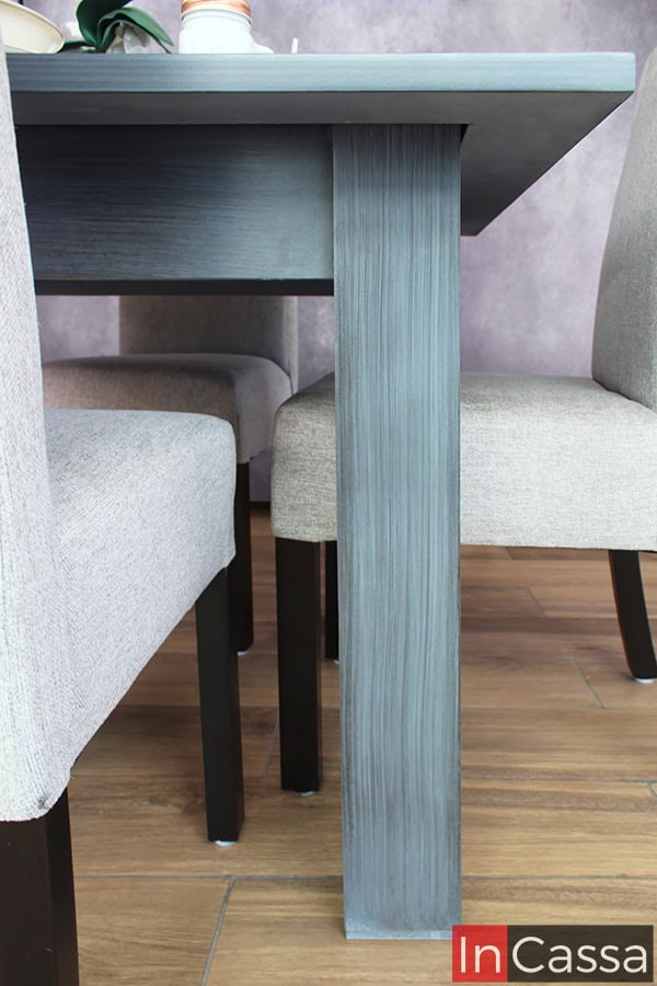 Una fotografía de una de las patas de la mesa en acabado gris cenizo oscuro, combinada con sillas tapizadas en tela gris claro. El diseño robusto y minimalista es perfecto para un comedor moderno.