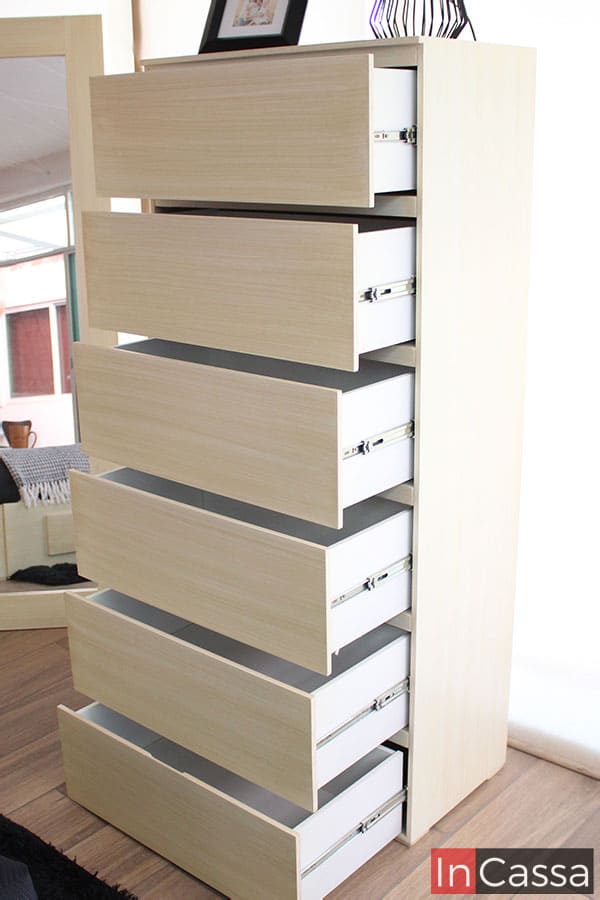 Una foto de la cajonera alta incluida en la recámara, la cuál posee 6 cajones amplios cuyo sistema de apertura y cierre consta de rieles metálicos. El diseño de cada cajón cuenta con un pequeño espacio en el borde superior, el cual permite la apertura de cada cajón sin necesidad de una jaladera. Todas y cada una de las parte que conforman el mueble conservan el mismo color paja.