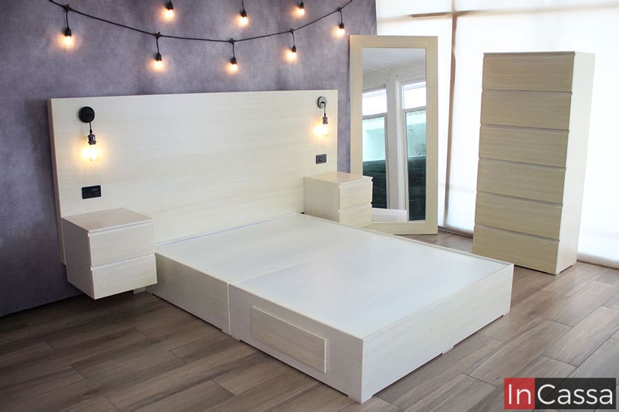 Se nos presenta la recámara completa, esta vez sin colchón en la base, dejándonos ver que la base se compone de dos partes. Además también vemos los buroes instalados en las contras integradas a la base, dándoles un moderno efecto flotante. La cabecera, buroes, espejo, cajonera y base comparten el mismo color paja.