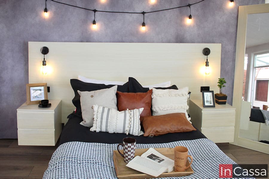 La imagen se enfoca en la cabecera y en los dos buroes de la recámara, los cuales son completamente de color paja; la cabecera cuenta con dos lamparas minimalistas integradas, además de que su diseño con contras de madera le permite sostener los dos buroes manteniéndolos suspendidos.