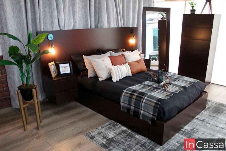 Moderna recámara matrimonial conformada por una cabecera con lamparas minimalistas integradas, dos buroes flotantes, un espejo de cuerpo completo y una cajonera, cada mueble del juego de recámara comparte el mismo tono tabaco. La recámara se encuentra instalada en una estancia con piso de madera y una pared cubierta por una cortina gris.