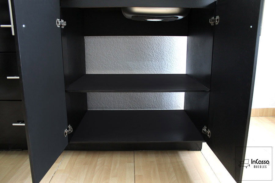 La foto presenta un gabinete inferior en color negro, ubicado debajo de la tarja, con dos puertas abatibles y bisagras metálicas. Su diseño incluye un estante interno que optimiza el almacenamiento de productos de limpieza o utensilios de cocina. La estructura es resistente y de fácil acceso.