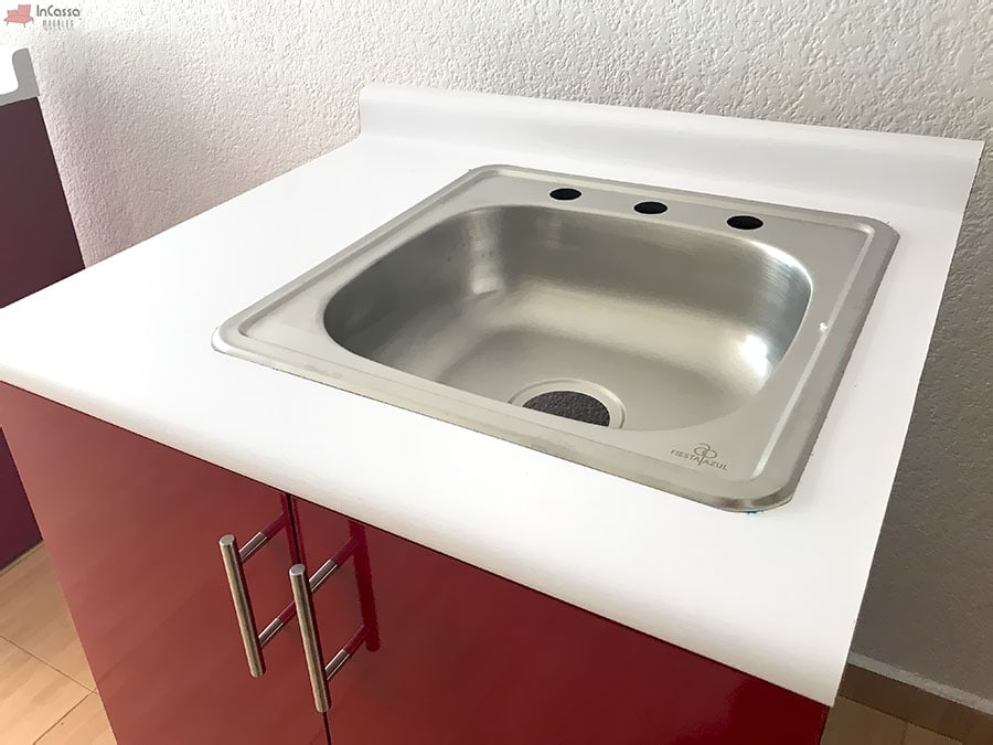 Una toma superior destaca la tarja de acero inoxidable con escurridor incluida en la cocina HAMPTON, empotrada sobre una cubierta de formica blanco fresh con respaldo. Ideal para garantizar higiene y durabilidad en el área de lavado.