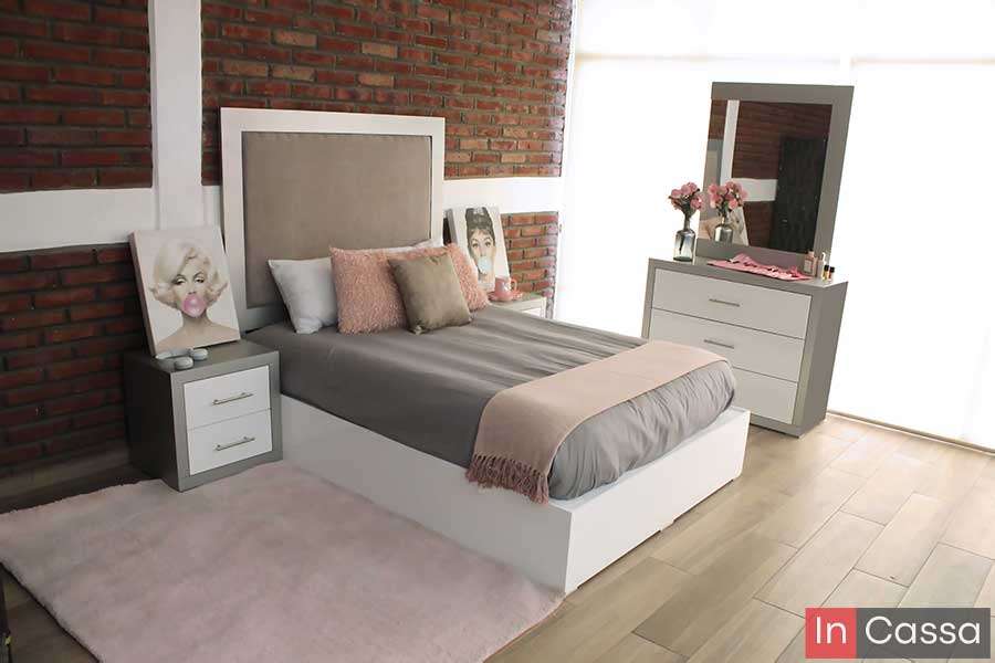 Una toma angular muestra una recámara moderna con cabecera tapizada, base blanca, buró con cajones y tocador con espejo amplio en color silver con cajones blancos. Los detalles en rosa y gris, junto con el muro de ladrillo, crean un ambiente acogedor y contemporáneo.
