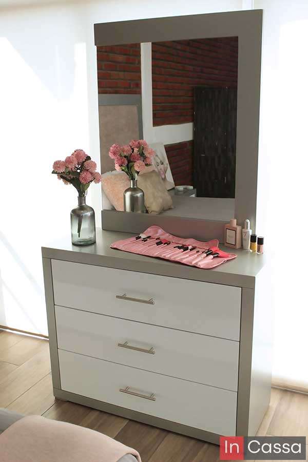 La imagen muestra el tocador de la recámara en color silver, con tres cajones blancos amplios, jaladeras metálicas y un espejo de marco grueso. Sobre la superficie destacan flores rosadas y un set de brochas, reflejando un estilo femenino, ordenado y contemporáneo.