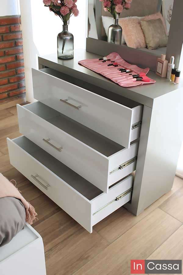 Una toma en ángulo muestra el tocador de la recámara con sus tres cajones abiertos, revelando su amplio espacio interior. El diseño bicolor silver y blanco con jaladeras metálicas y espejo superior lo convierte en una pieza práctica y estética para cualquier recámara.