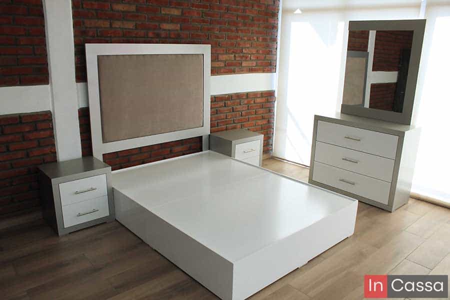 Una toma angular presenta la recámara sin colchón, destacando la base blanca de líneas rectas, cabecera tapizada en suede camel y dos burós silver con blanco. A la derecha, el tocador con espejo completa este conjunto moderno y funcional.